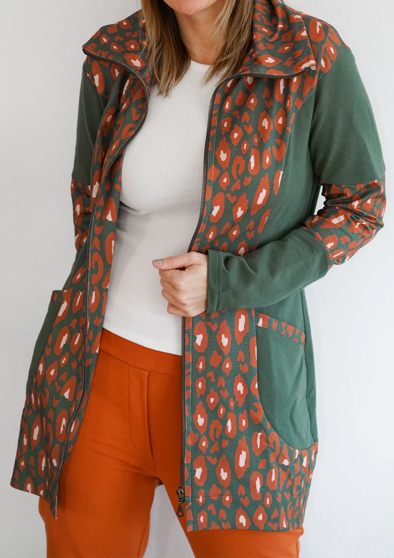 Woman wearing a green jacket with orange leopard-print panels, a white knit top, and orange pants; the zipper is halfway up on a plain background.